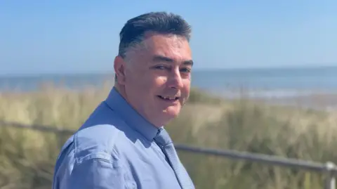 A man in a blue shirt is standing in front of the sand dunes on a sunny day. He has black hair, swept back. The sea can be seen under a blue sky.