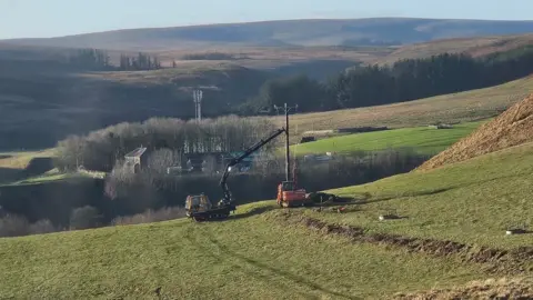 Shona Anderson A pole is being put up by a truck with a long extendible arm which is holding it in place. In the distance there is a farm and a telecommunications mast, while desolate hills poke up in the distance.