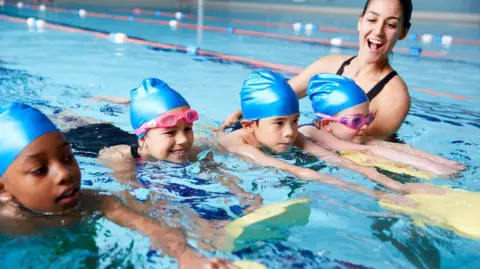 A swimming coach teaching four children how to swim. The children are wearing blue swimming caps and holding yellow floats.