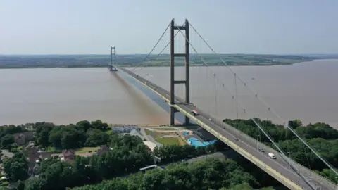 The Humber Bridge stretching across the Humber. The bridge has tall towers and cables supporting it, with vehicles traveling in both directions. Green countryside and scattered houses can be seen on either side of the Humber. The sky is clear and blue.