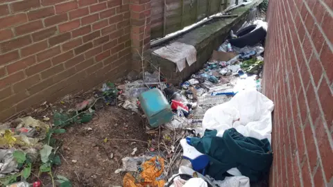 Peterborough Litter Wombles Rubbish dumped in a narrow brick wall-lined alleyway. It shows clothes, scattered tin cans, cardboard, a metal rack, a tyre, plastic bags. 