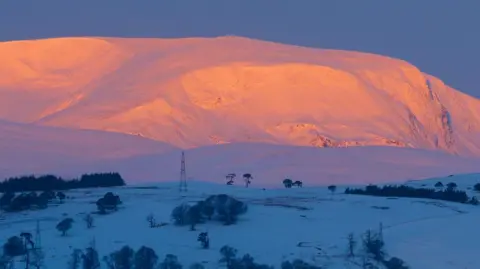 Dominic McArdle Snow-covered Ben Wyvis mountain glows pink and orange in the light of the sunrise. Fields in the foreground are in shadow and a chilly blue colour.