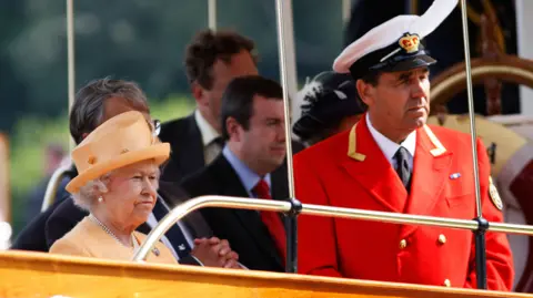 Getty Images Queen Elizabeth II, wearing an orange outfit and hat, stands next to David Barber, who is wearing a smart red coat and naval-style cap bearing a crown emblem.