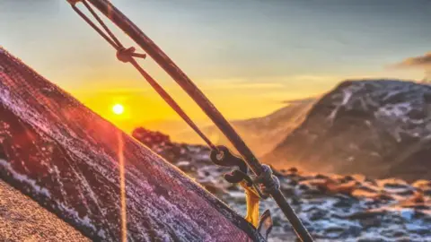 Alan O’Connor A frosty tent at the top of a mountain range with a yellow/orange sunrise in the distance, next to snow-covered peaks.