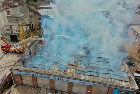 Stephen Huntley/BBC Two firefighters on an aerial platform spraying water over the what is left of the roof of a building. Only the blackened frame of the roof remains and smoke is rising from it. There is an industrial building to the left. 