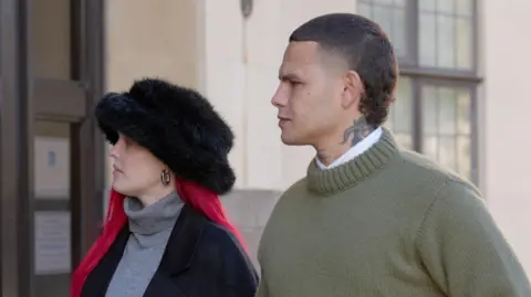 Tyron Frampton (right), known as Slowthai, walking outside a court wearing a green jumper. He has a tattoo on his neck and a short mullet haircut. A woman with long red hair and a furry hat walks beside him.