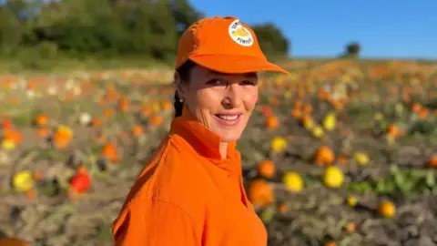 George Carden/BBC Farmer Caroline Harriott in a pumpkin field wearing an orange had and top.
