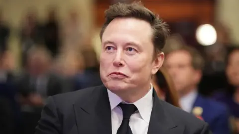 Getty Images Elon Musk, with dark hair wearing a black suit and tie with a white shirt, looks to his right while sat in an audience with blurred out people behind him 