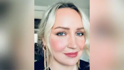 RIP.IE A woman is sitting in a living room. She's smiling into he camera. She has blond hair and is wearing gold earrings.