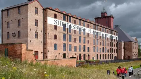 The front of a large, five storey brick building with two towers and a small group of children in red tops walking near the base. It has the words Shrewsbury Flaxmill Maltings written on a large white sign on the wall.