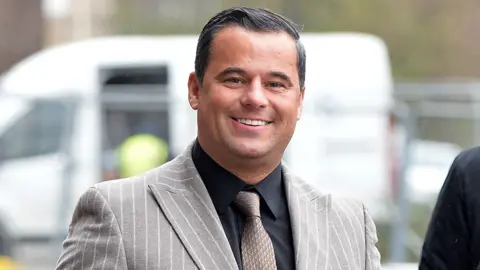 James English wearing a grey and white striped suit, black shirt and brown tie. He is walking into court, smiling at the camera.