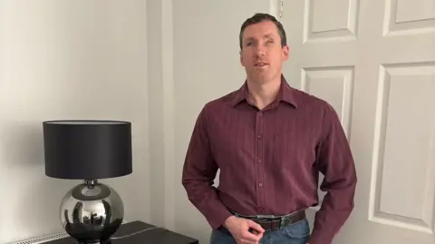 Liam Harkness Liam Harkness is pictured standing in front of a white door wearing a burgundy shirt and blue jeans. One of his hands is resting on his belt. Next to him is a metallic table lamp with a black shade. 