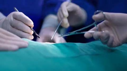 Photo of hands of operating room staff performing surgery. The instruments being held include a scalpel and forceps. There is a green medical sheet covering the patient.
