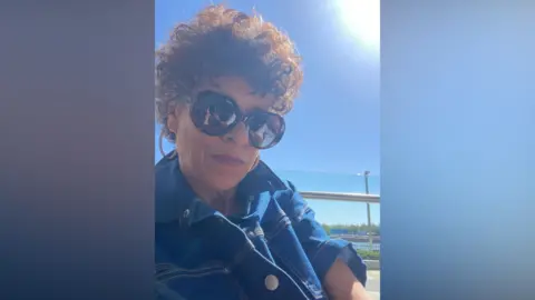 Carolyn Stewart/Instagram A lady with curly brown hair and large sunglasses is pictured on a sunny day with a blue sky behind her. She is wearing a blue denim jacket.