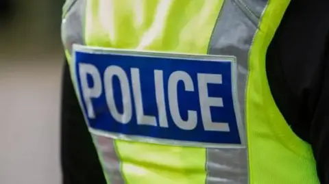 A person wearing a hi-vis gilet and black top. In the centre of the gilet is the word Police in white lettering on a blue background. 