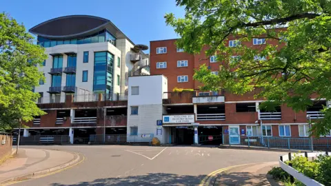 A car park. The car park is made of red and white bricks. A road bordered by trees sits in front of the car park.