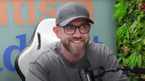 Andrew Williams A man in a grey top and grey baseball cap with a brown beard, sitting behind a black microphone in front of a grey wall with plants to his left hand side