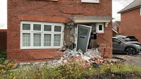 The front door of a house smashed in with bricks and rubble lying on the grass 