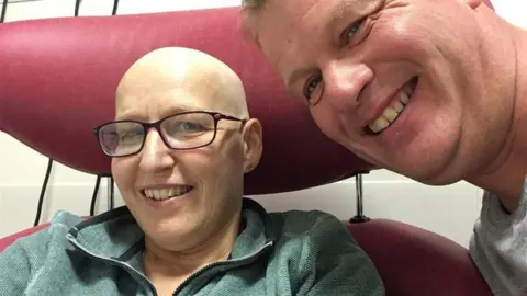 A woman sits in a hospital chair, smiling in a selfie while her husband leans his head towards her. She has no hair and is wearing glasses.