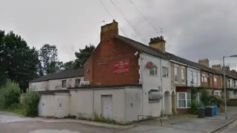 Google A Google Streetview of the side of a terrace building at the end of a street. It says "New Delphi Club" in yellow writing with a red background on a brick wall. There are terrace houses next to it.