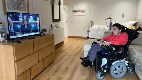 A woman sitting in a motorised wheelchair, she is watching the tv. She is wearing a red jumper and red and white check shirt