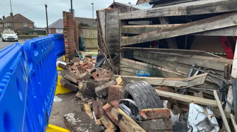 George Carden/BBC Bricks still scattered on the floor following the incident on 30 January. One of the car's wheels and part of its bumper are still lodged among the debris. A blue plastic fence is set up on the left to cordon off the debris from the road and part of a wooden shed is also damaged. 