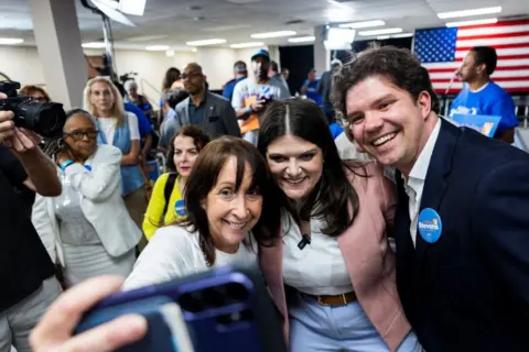 Getty Images Three people are taking a selfie in a crowded room. On the left of the three is a middle aged woman holding the camera. In the middle is Haley Stevens with a broad smile. On the right is Gary Peters dressed in a dark suit and open necked, white shirt.