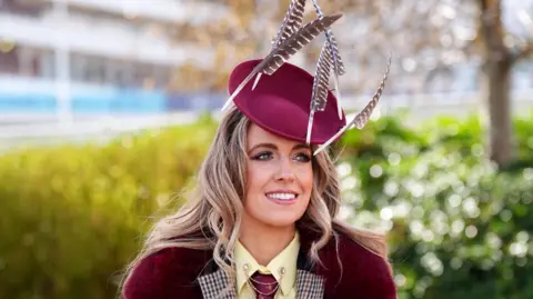 PA Media A young woman with wavy blonde hair smiles as she poses for the camera at Ladies Day at the Cheltenham Festival 2026. She is wearing a plum red berry-style beret with feathers attached to it, and a jacket that matches her hat