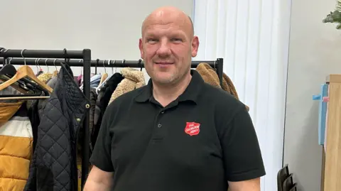 John Bullock stood in front of a rail of donated coats. He is wearing a black t-shirt which has a red badge on it which says "The Salvation Army". 