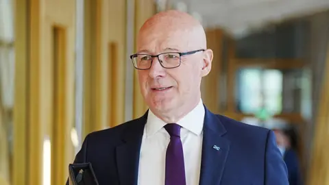 PA Media John Swinney wearing a blue blazer, purple tie, white shirt and glasses. He is walking down a corridor at Holyrood.