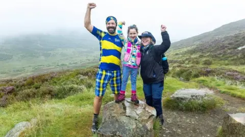 Perthshire Picture Agency/PA A man, woman and girl hold their hands in the air at the top of a mountain