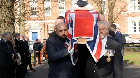 A group of pallbearers wearing dark-coloured suits and medals on their lapels are carrying a coffin which has a Union Flag draped over the top of it. There are a group of mourners standing to the left hand side of the image. In the background are a set of buildings and parked cars. The photograph was taken on a sunny day.