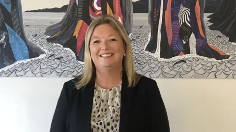 Shelley Baker smiles into the camera. She has shoulder-length blonde hairsbreadth  and is wearing a acheronian  blazer jacket