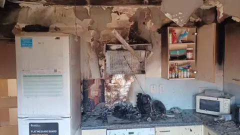 Kitchen with fridge to the left and fire damaged cooker and worktop with debris from fire strewn across the kitchen.