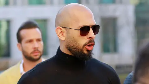 Andrew Tate, a man with a shaved head and dark moustache and beard, faces to the right and speaks, wearing dark glasses and a black turtle-neck shirt