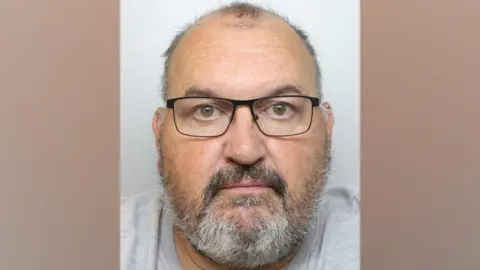 A police custody photograph of Allan Wood who has short grey hair, a grey beard and is wearing dark framed glasses and a grey t-shirt