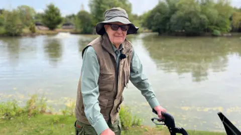 BBC Peter Byers is stood with his rollator walking aid in front of a lake. He is wearing a green fishing hat, sunglasses, a khaki fishing gilet and a grey-green shirt.