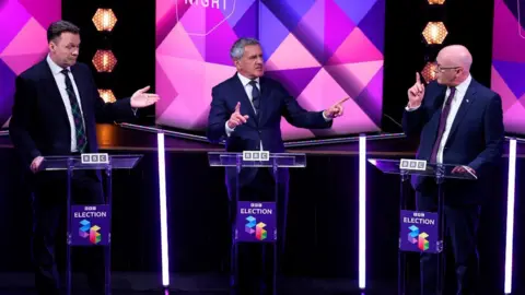Three men in business suits stand at podium gesticulating at each other 