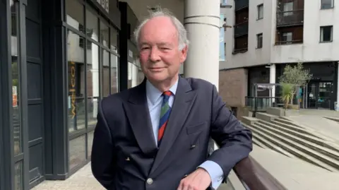 Mr Seccombe is stood outside some buildings wearing a navy suit and blue, green and red tie with a light blue shirt on underneath. He is leaning on a railing smiling at the camera.