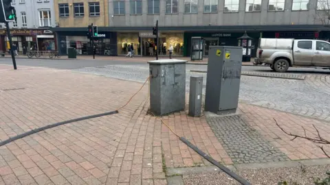 A grey electricity box on the pavement of a street in Taunton. In the background is a road and shops and a bank.