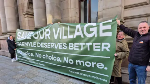 A group of people hold up a very large banner outside the City Chambers. It has white letters on a green background saying SAVE OUR VILLAGE - CARMYLE DESERVES BETTER. It also says No voice. No choice. No more underneath the main text.