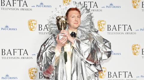 Comedian Joe Lycett dressed as Queen Elizabeth I in a silver gown with a large collar. He is holding a Bafta award
