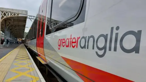 A white, red and grey Greater Anglia train with its doors closed at a railway station.