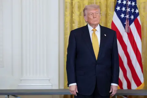 Donald Trump, in a dark navy suit and yellow tie, stands with his hands at his sides and a neutral expression. Behind him are gold curtains in the White House and an American flag. 