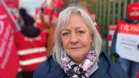 A woman in a navy coat looks into the camera. Behind her, out-of-focus is a group of people in red vests holding flags and placards. The woman wears a pink, white and blue handkerchief. She has long grey/white hair.