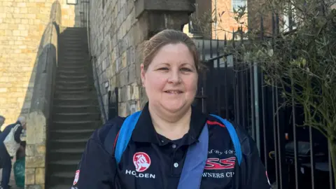 BBC/ Emily Johnson A woman, with tied up blonde hair in a sports top, smiles in front of a stone staircase leading up to York's city walls.