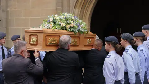PA Media A light brown wooden coffin is carried on shoulders inside a doorway, young people in light blue shirts wearing berets stand guard either side of the coffin.