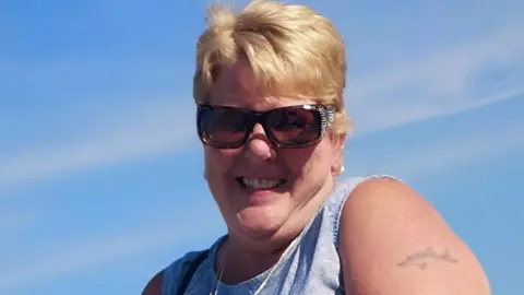 Family handout A woman with short blonde hair smiles at the camera against a backdrop of blue sky. She is wearing sunglasses.