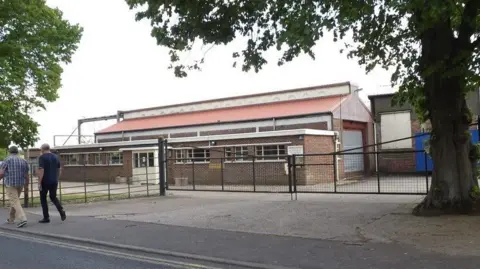 A large factory building can be seen with two men walking past towards the left along the pavement next to the building, which is located behind fencing and a gate. 