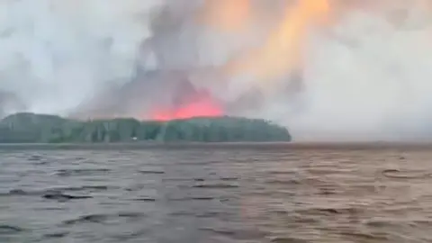 Smoke and flare behind a forest, seen from afar in the ocean.